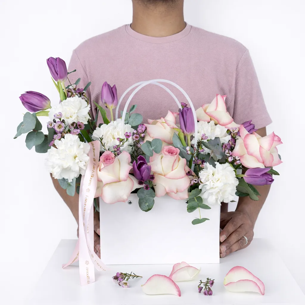 Person holding a white handled bag filled with purple tulips, cream roses with pink edges, white carnations, and pink wax flowers with eucalyptus and a Favish Blooms ribbon.