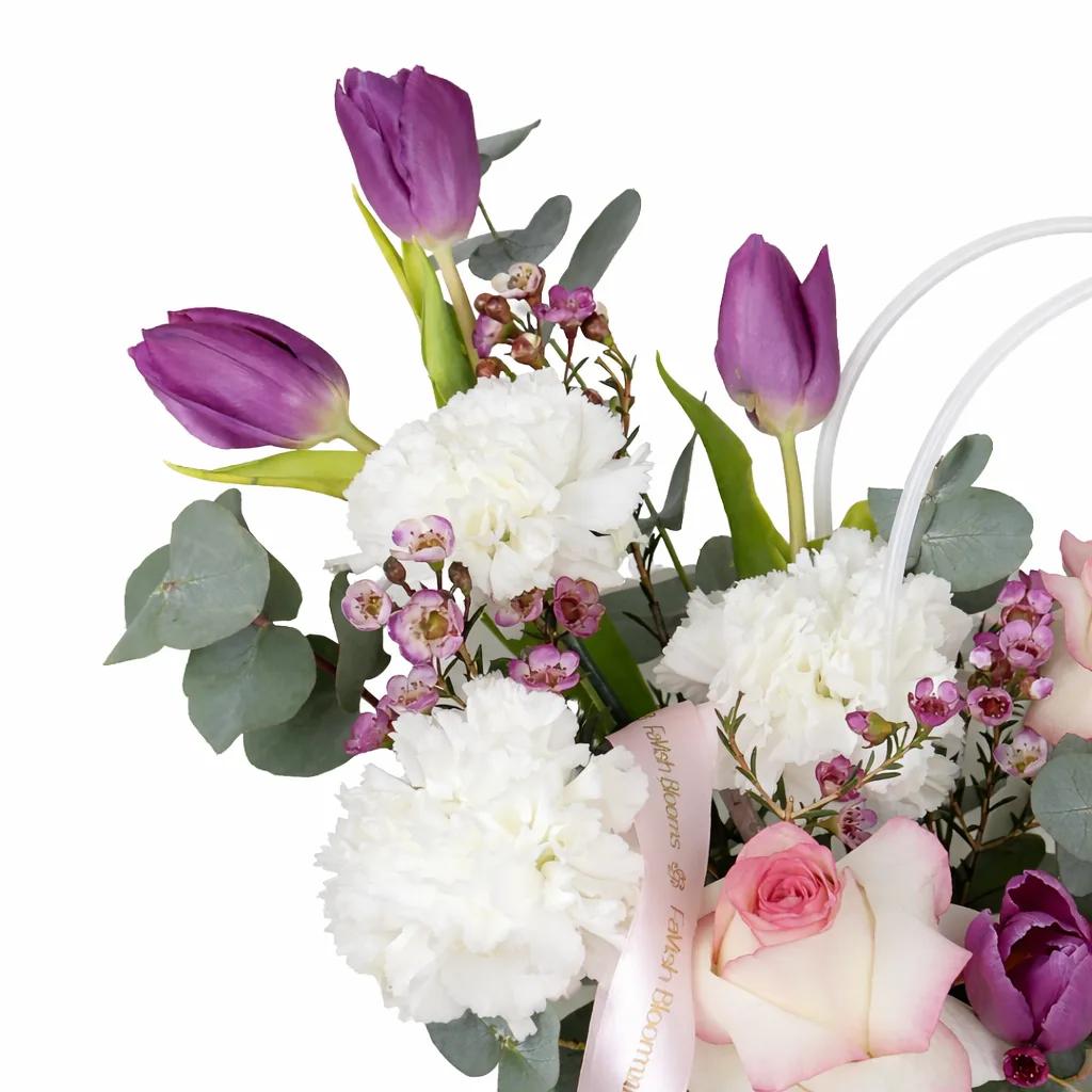 Close-up of purple tulips, white carnations, pink roses, and pink wax flowers with eucalyptus featuring a Favish Blooms ribbon.