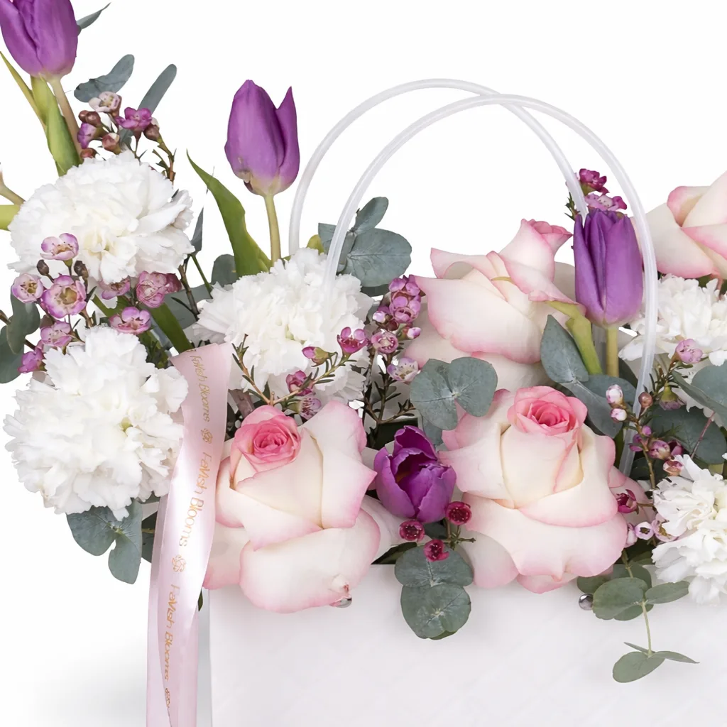Close-up of cream roses, purple tulips, white carnations, and pink wax flowers with eucalyptus in a white handled bag with a Favish Blooms ribbon.