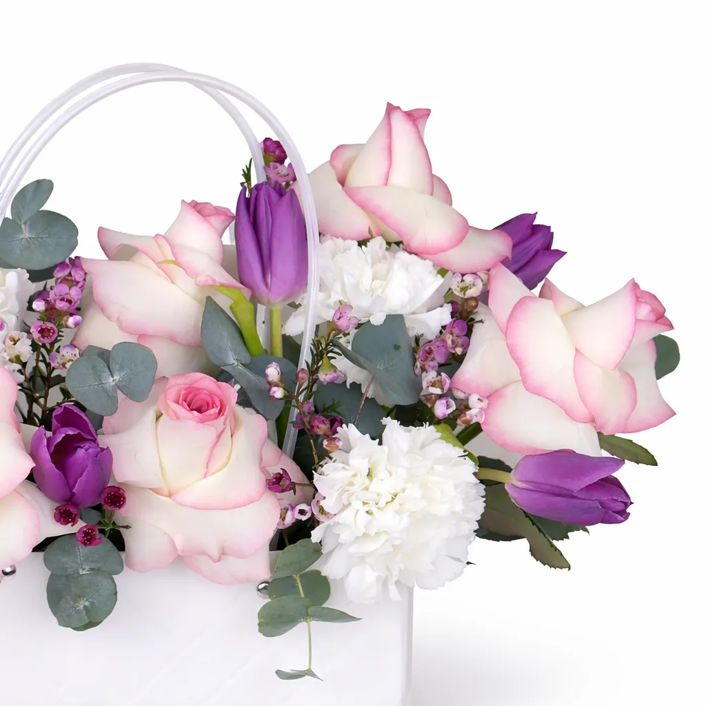Macro shot of cream roses with pink edges, purple tulips, white carnations, and pink wax flowers with eucalyptus in a white handled bag.
