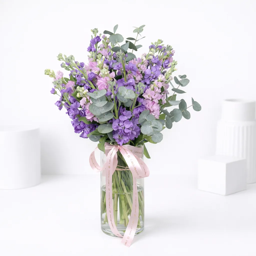 Purple and pink stock flowers with eucalyptus in a glass vase tied with a pink ribbon.