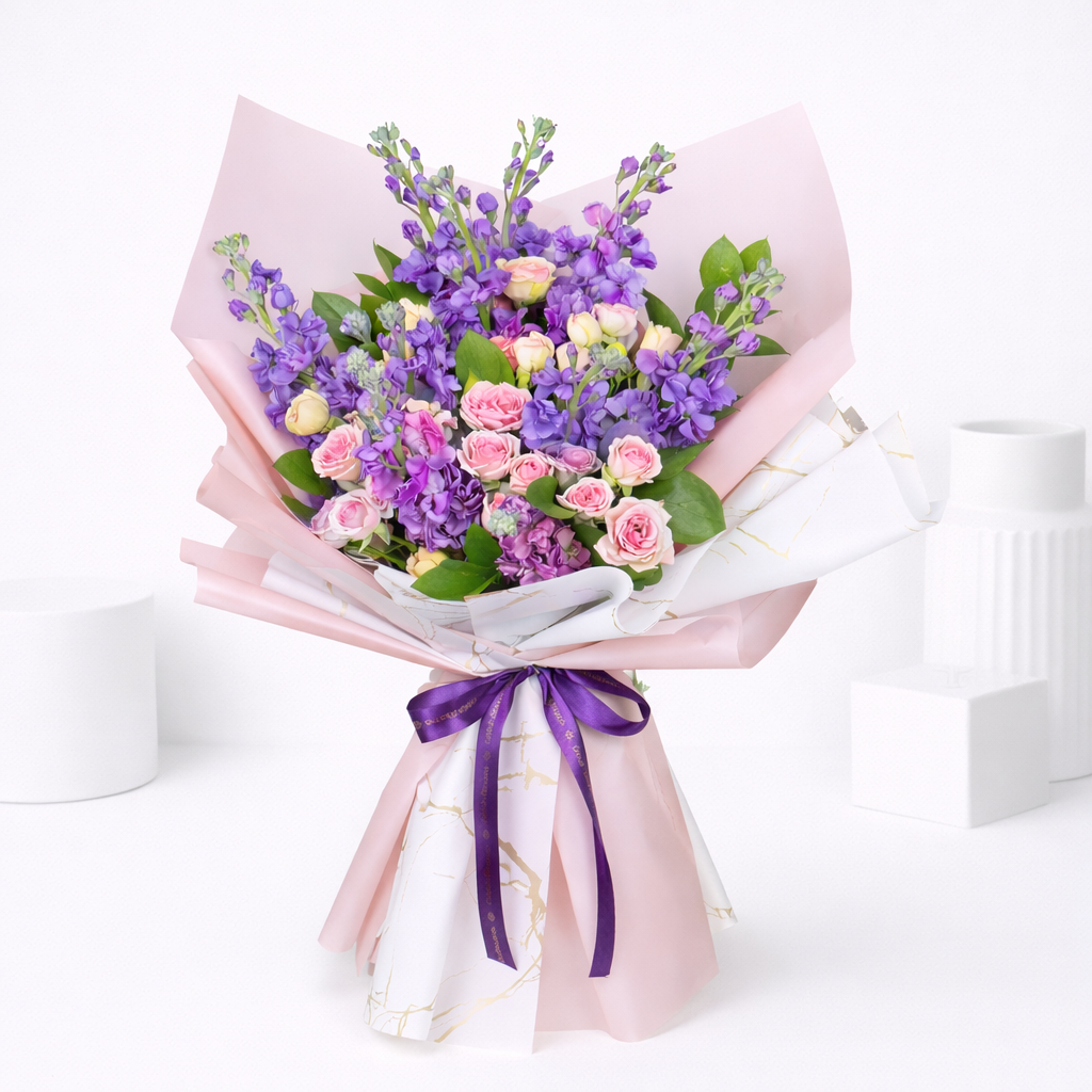 Purple stock flowers and pink spray roses bouquet wrapped in pink and marble paper with purple ribbon.