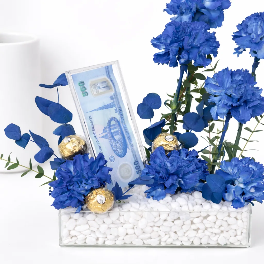Close-up of a blue carnation gift arrangement with Ferrero Rocher chocolates and a UAE 500 Dirham note in a clear acrylic tray.