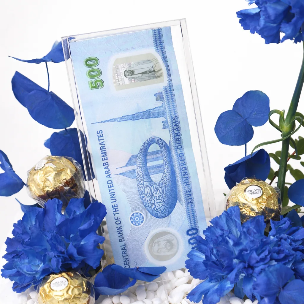 Close-up of a UAE 500 Dirham note in an acrylic holder surrounded by blue carnations and Ferrero Rocher chocolates.