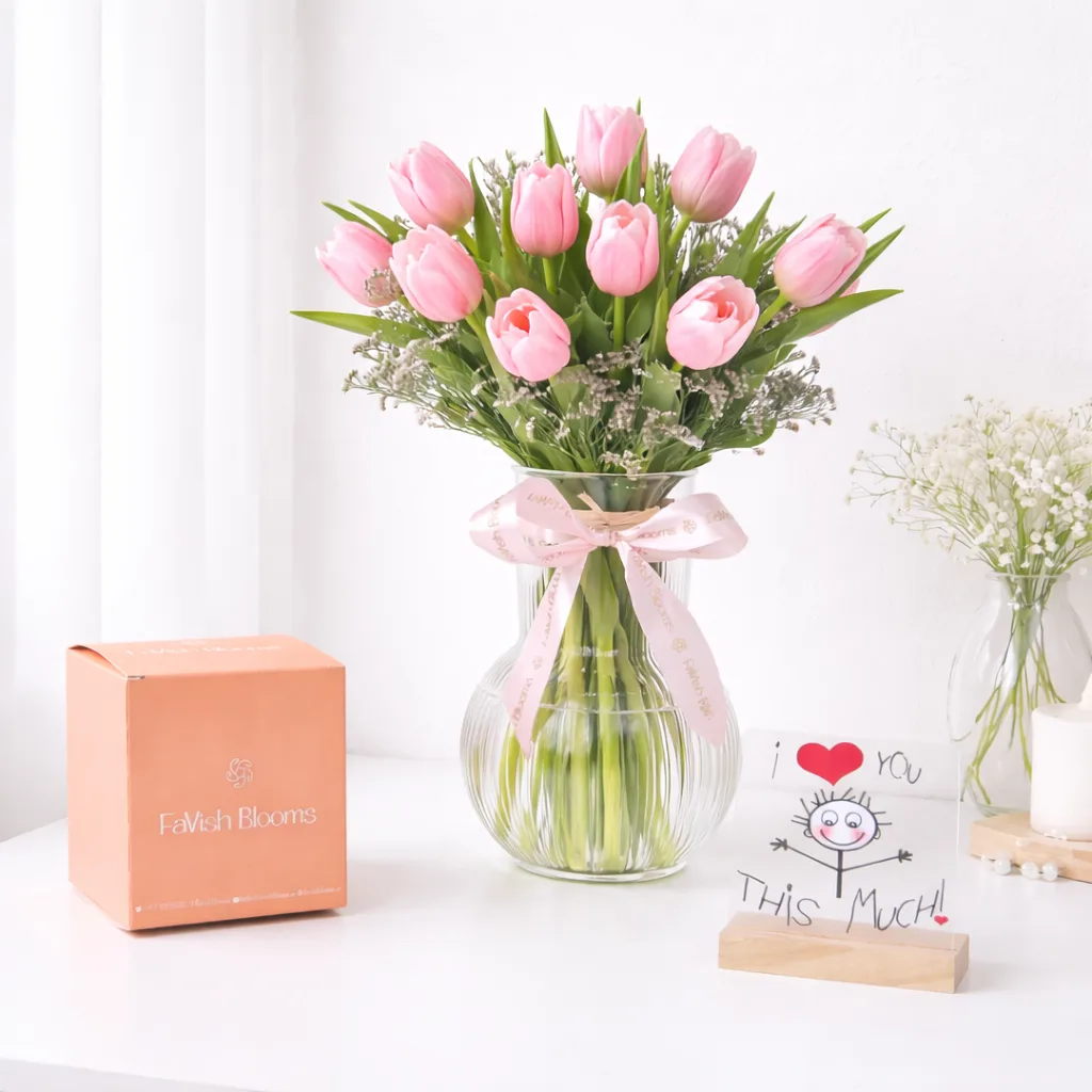 Pink tulip bouquet in a glass vase with a pink ribbon, FaVish Blooms gift box, and an 'I Love You This Much' greeting card on a white table.