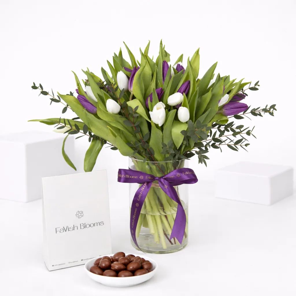 Purple and white tulips in a glass vase with a purple ribbon, beside chocolate-covered nuts and a FaVish Blooms card.