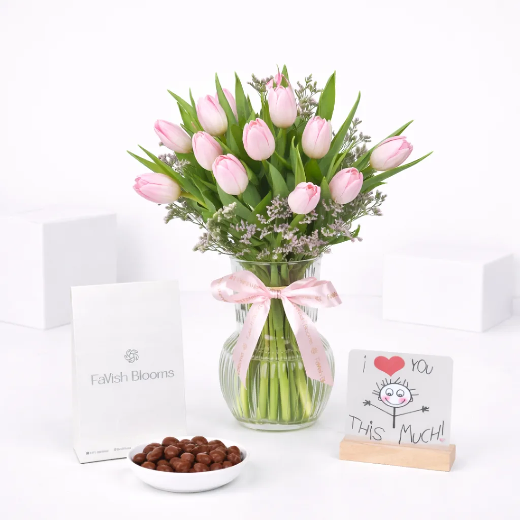 Pink tulip bouquet in a glass vase with a pink ribbon, FaVish Blooms bag, chocolate balls, and an 'I Love You This Much' greeting card on a white surface.