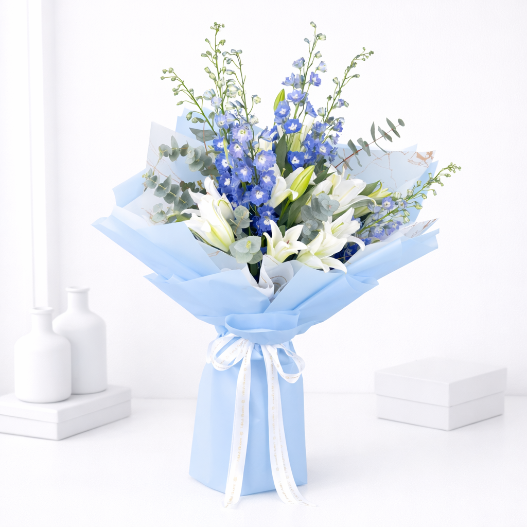 White lilies and blue delphinium bouquet wrapped in light blue paper with a white ribbon bow on a white background.
