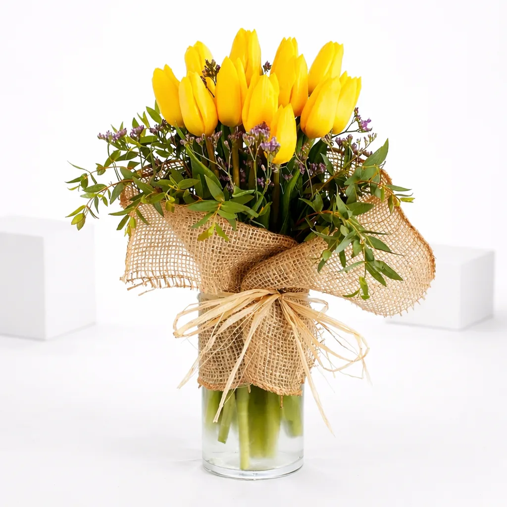 Yellow tulip bouquet with greenery in a glass vase wrapped in burlap and raffia bow.