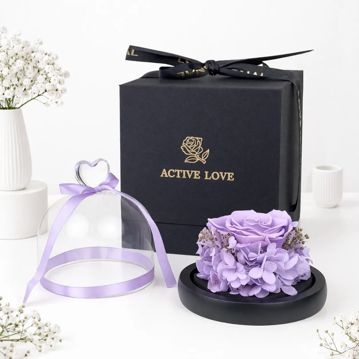 Purple preserved rose and hydrangea arrangement on a black base with a glass dome and ribbon, beside an "Active Love" black gift box.