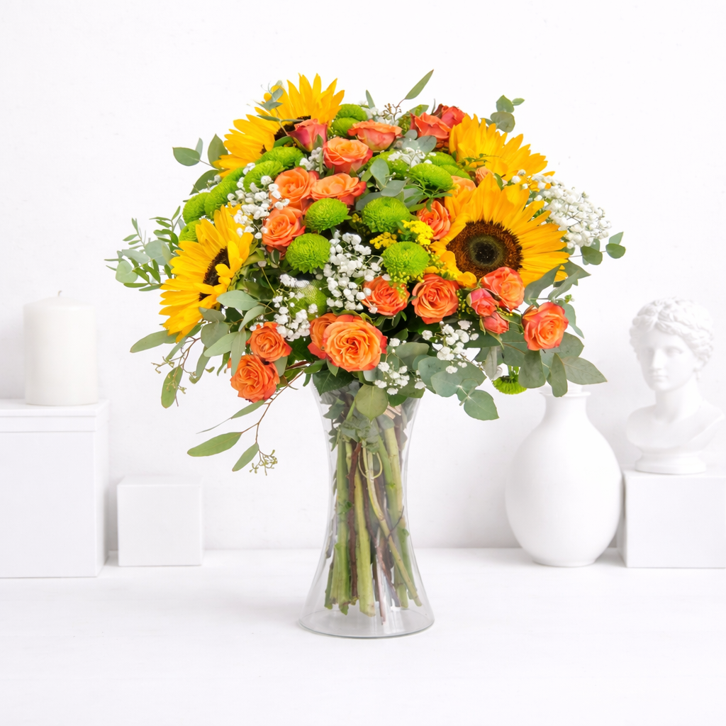 Vibrant sunflower and orange rose bouquet in a clear glass vase on a white surface.