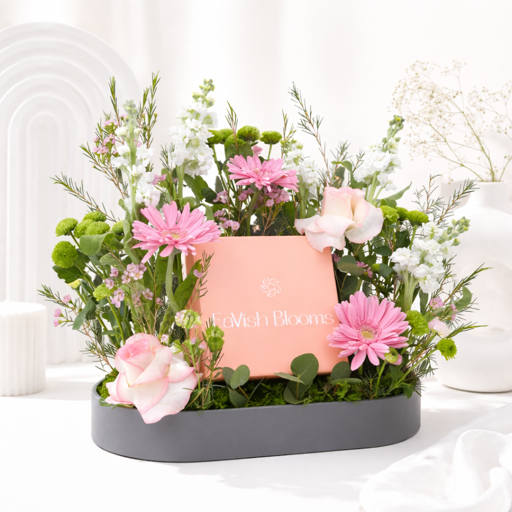 A floral arrangement of pink gerberas, roses, white blooms, and green button mums surrounding a pink gift box in a grey tray.