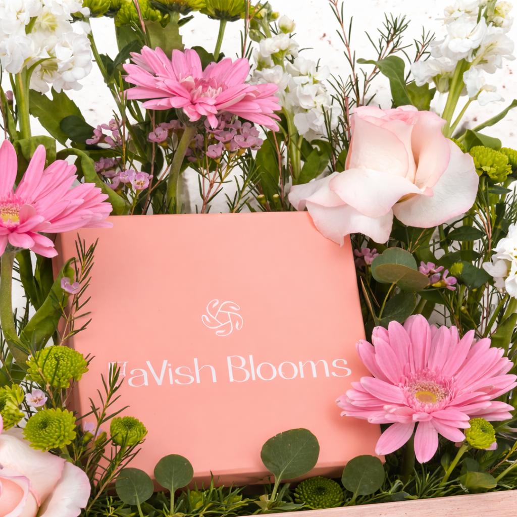 Close-up of a pink gift box surrounded by pink gerberas, roses, white blooms, and green button mums.