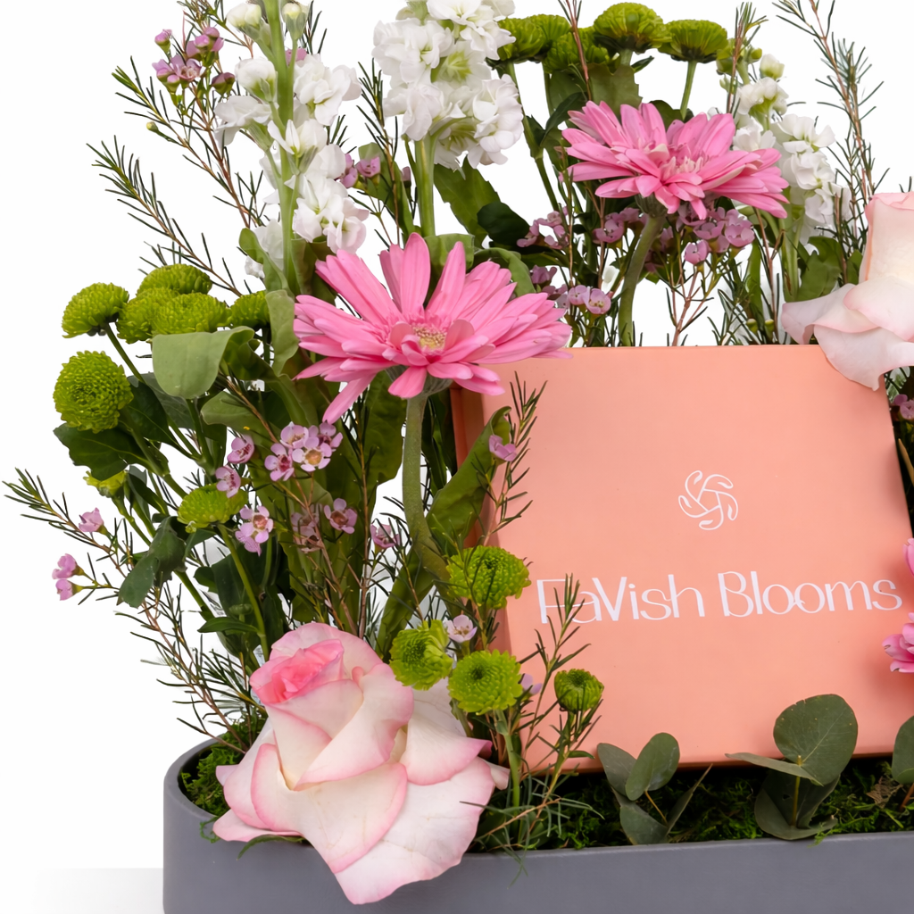 Close-up of pink gerberas, roses, white stock flowers, and green button mums around a pink gift box in a grey tray.