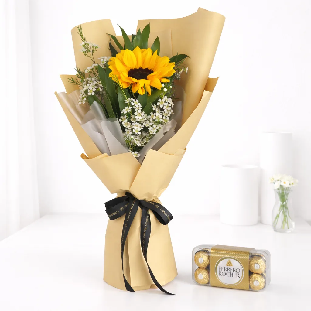 A sunflower bouquet with white filler flowers wrapped in kraft paper tied with a black ribbon, alongside a box of Ferrero Rocher chocolates.