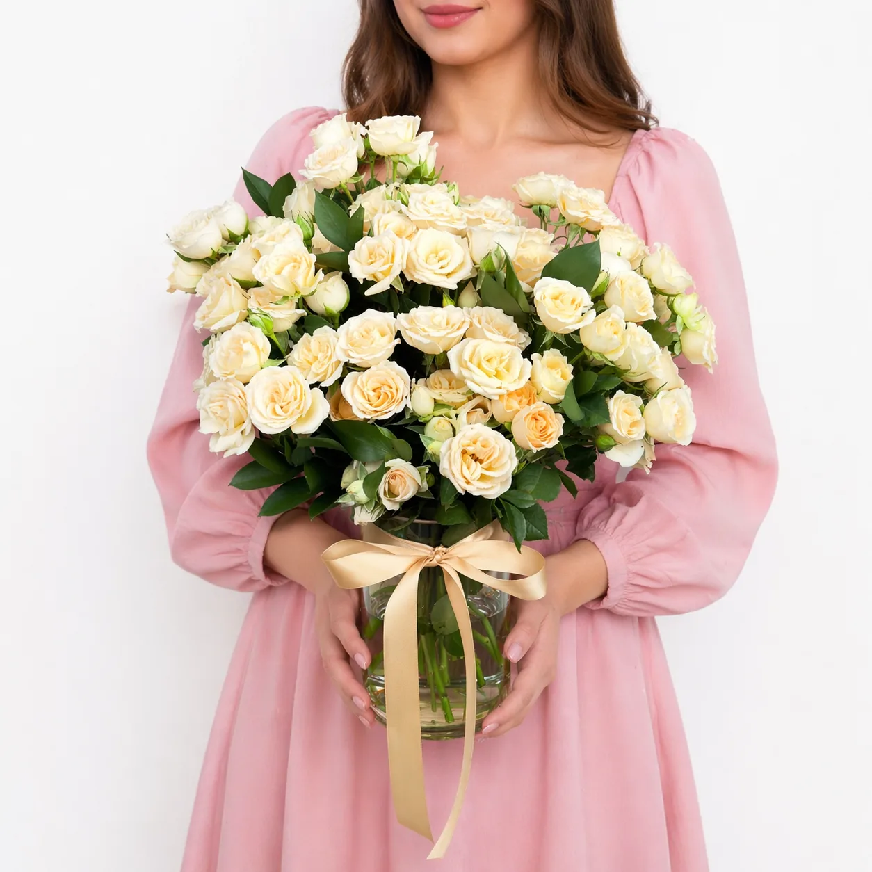 Woman in a pink dress holding a glass vase of cream spray roses tied with a champagne satin ribbon.