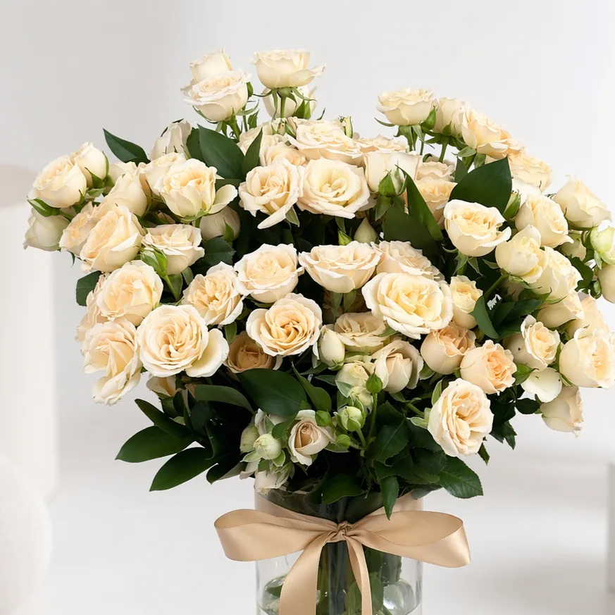 Close-up of a lush bouquet of cream spray roses in a glass vase tied with a champagne satin ribbon.