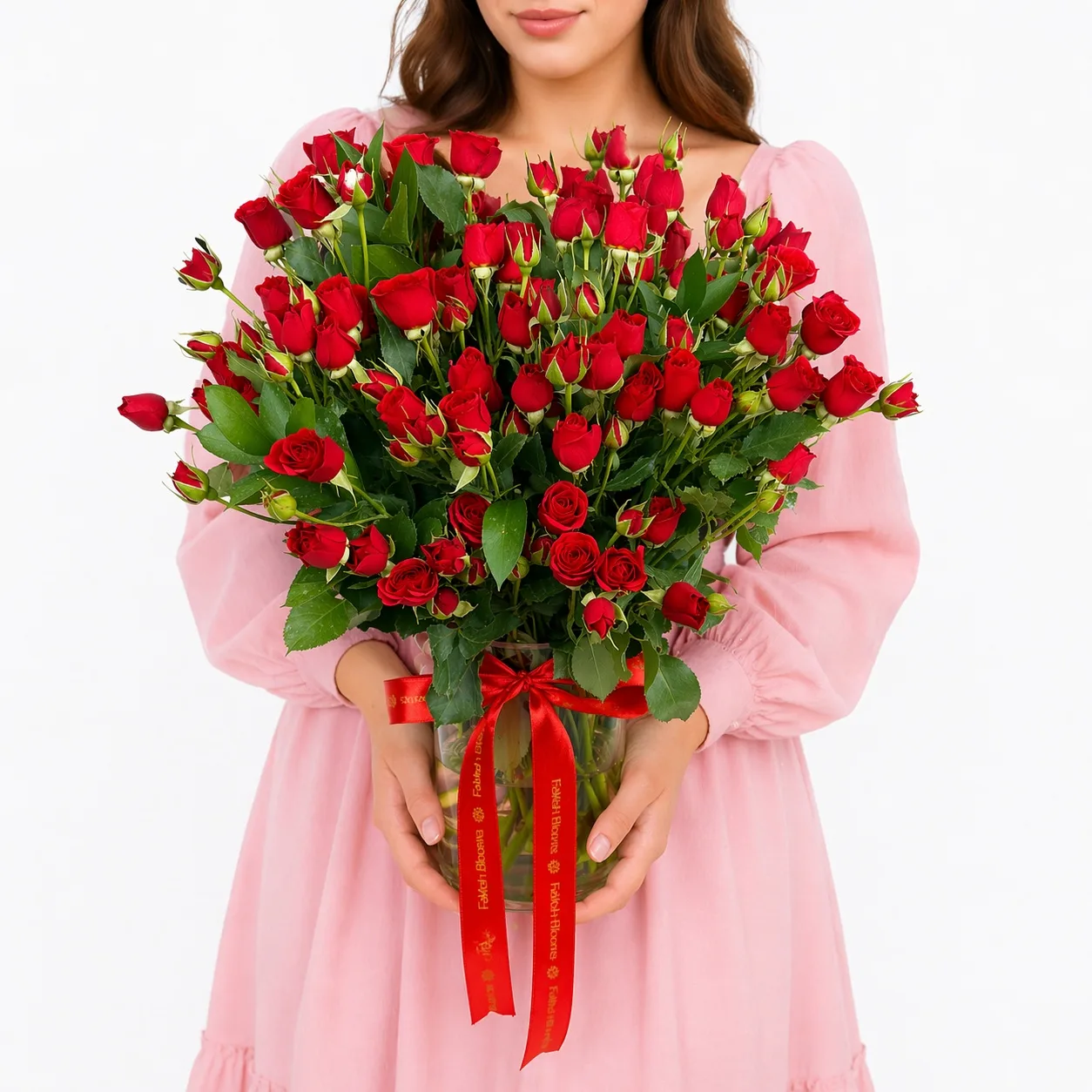 Woman in a pink dress holding a glass vase of red spray roses tied with a red satin ribbon.