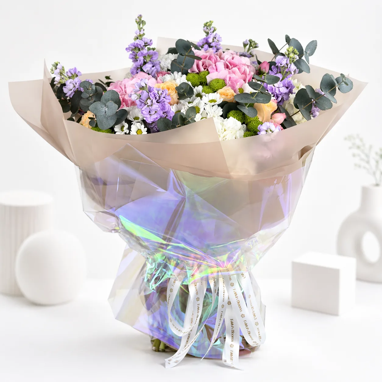 Pastel bouquet of pink hydrangeas, peach roses, purple stock flowers, white daisies, and eucalyptus wrapped in kraft paper and iridescent cellophane.