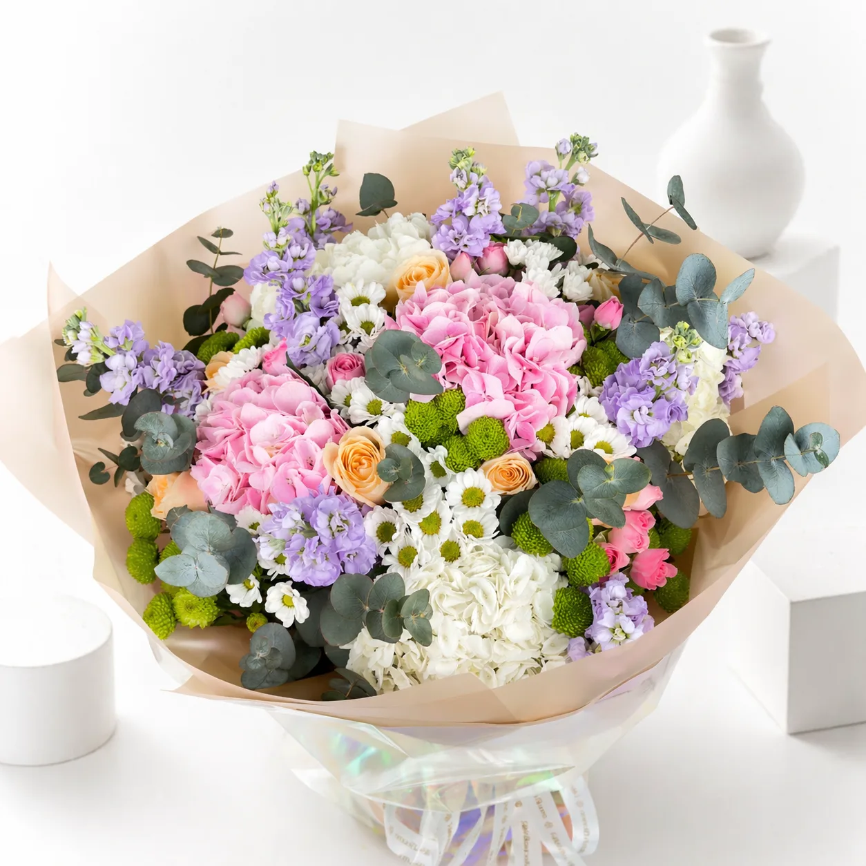 Large pastel bouquet of pink and white hydrangeas, peach roses, purple stock flowers, white daisies, and eucalyptus wrapped in kraft paper.