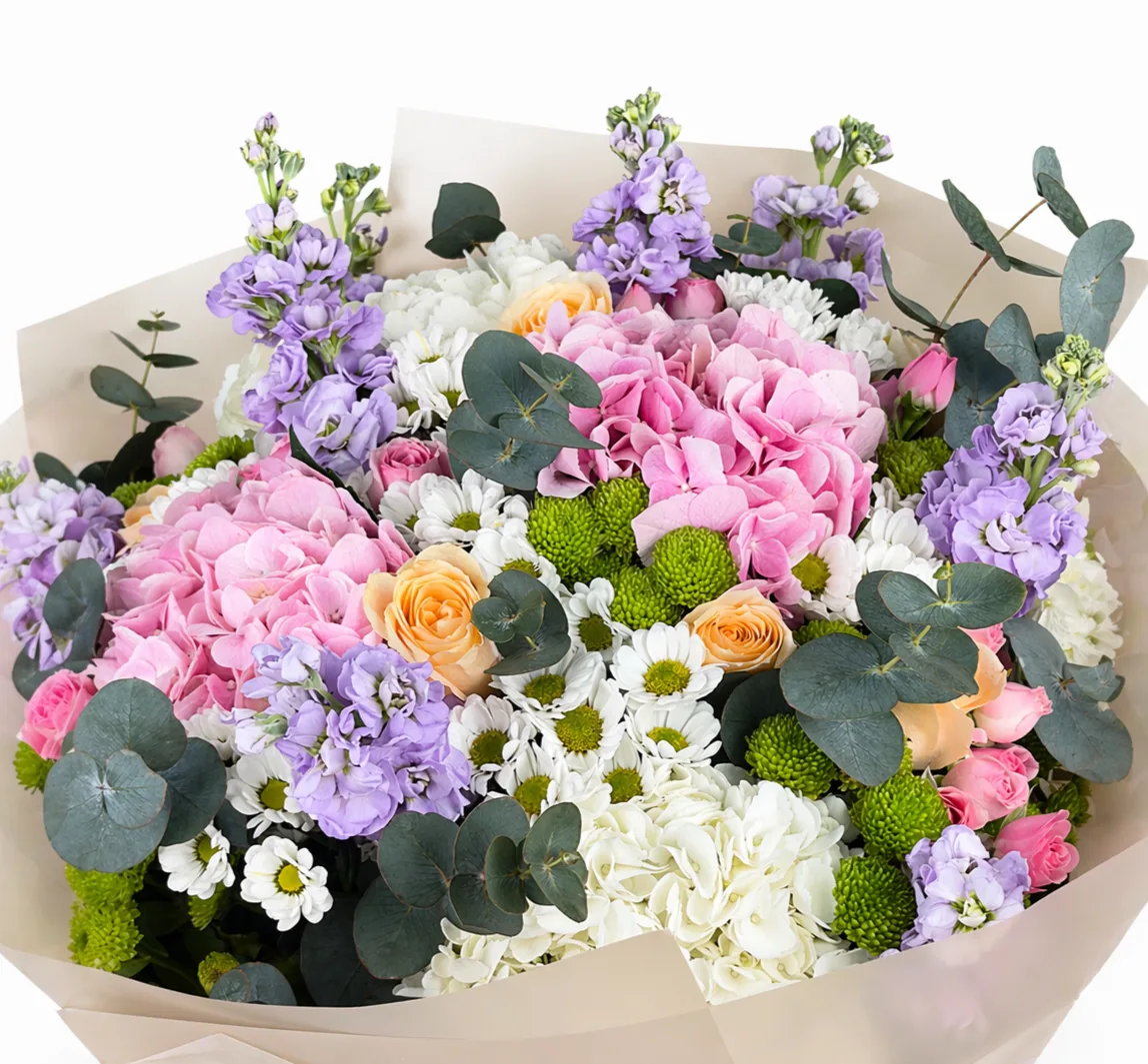 Close-up of a pastel bouquet featuring pink and white hydrangeas, peach roses, purple stock flowers, white daisies, green chrysanthemums, and eucalyptus in kraft paper wrapping
