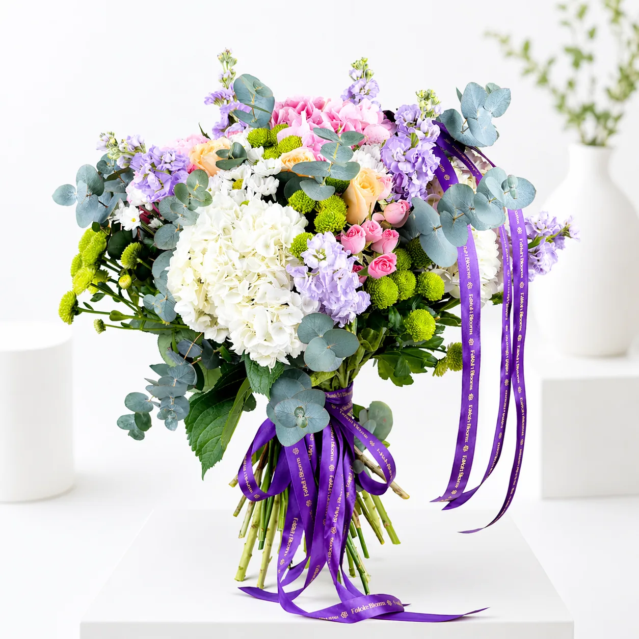 Pastel bouquet of white hydrangeas, pink roses, purple stock flowers, green chrysanthemums, and eucalyptus tied with a purple satin ribbon.