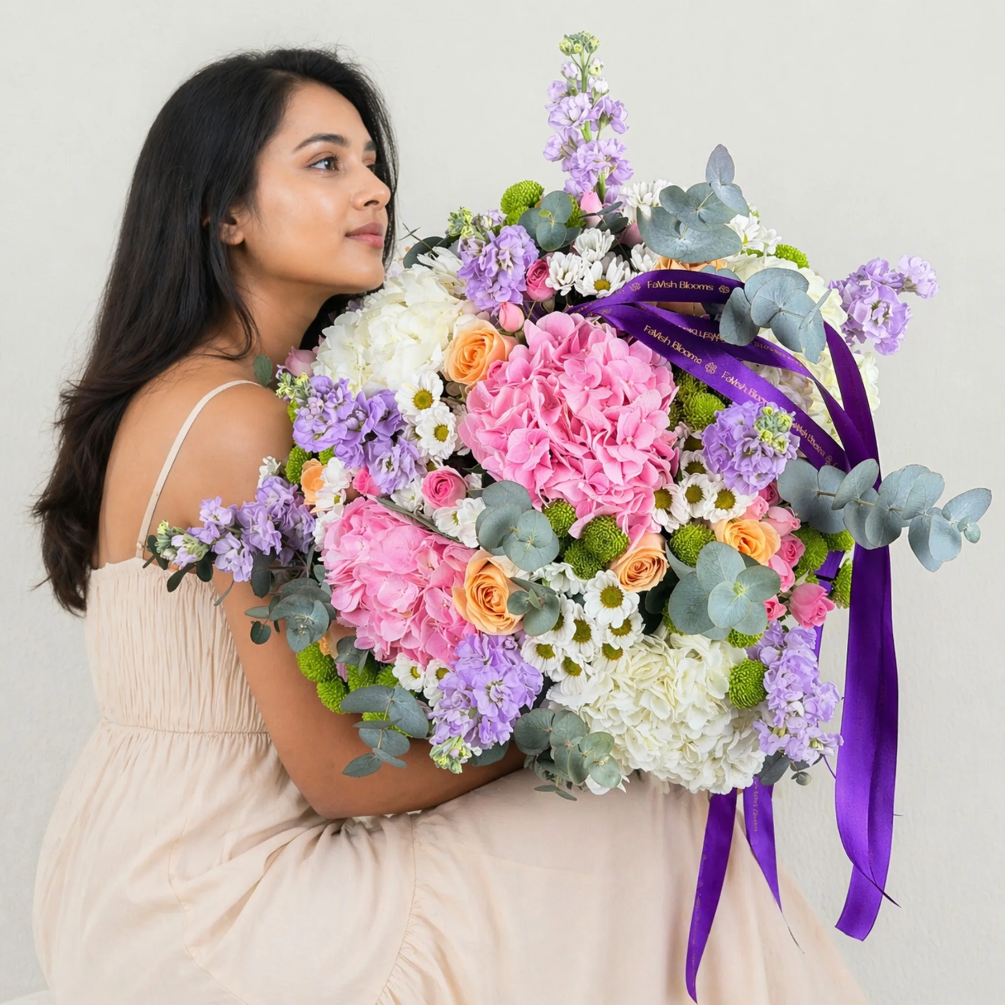Woman in a beige dress holding a large pastel bouquet of pink and white hydrangeas, peach roses, purple stock flowers, white daisies, and eucalyptus tied with a purple ribbon.
