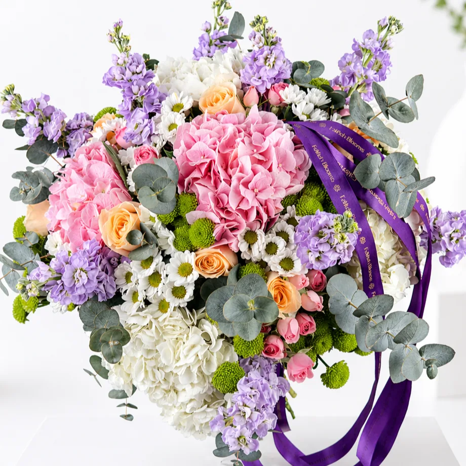 Close-up of a pastel bouquet with pink and white hydrangeas, peach roses, purple stock flowers, white daisies, and eucalyptus tied with a purple ribbon.