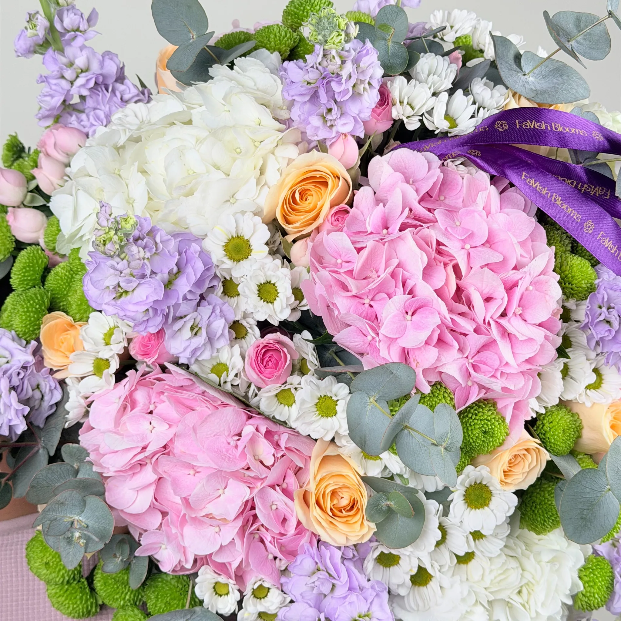 Macro shot of pink and white hydrangeas, peach roses, purple stock flowers, white daisies, and green chrysanthemums with a purple branded ribbon.
