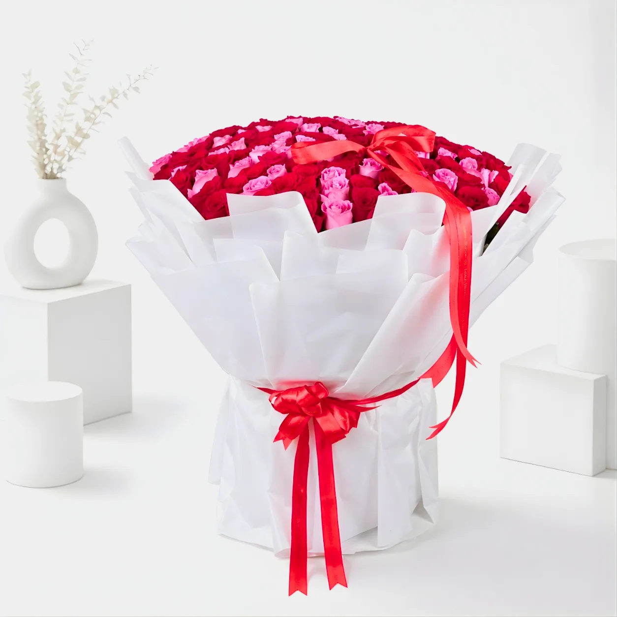 Luxury red and pink rose bouquet wrapped in white paper with red ribbon, styled in a bright white studio setup.