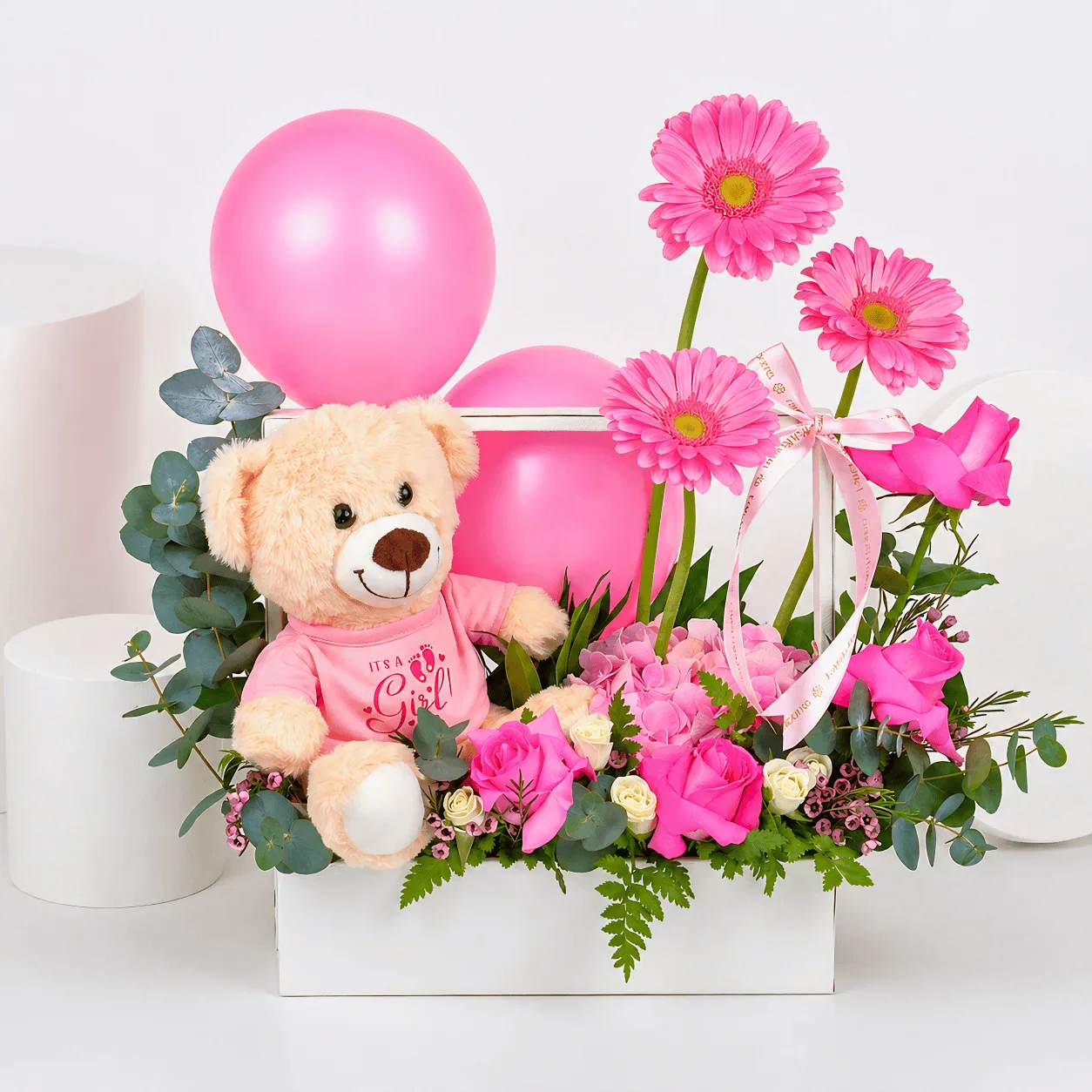 Pink baby girl gift arrangement with teddy bear, balloons, gerbera flowers, pink roses, hydrangea, eucalyptus, and fresh greenery in a white box.