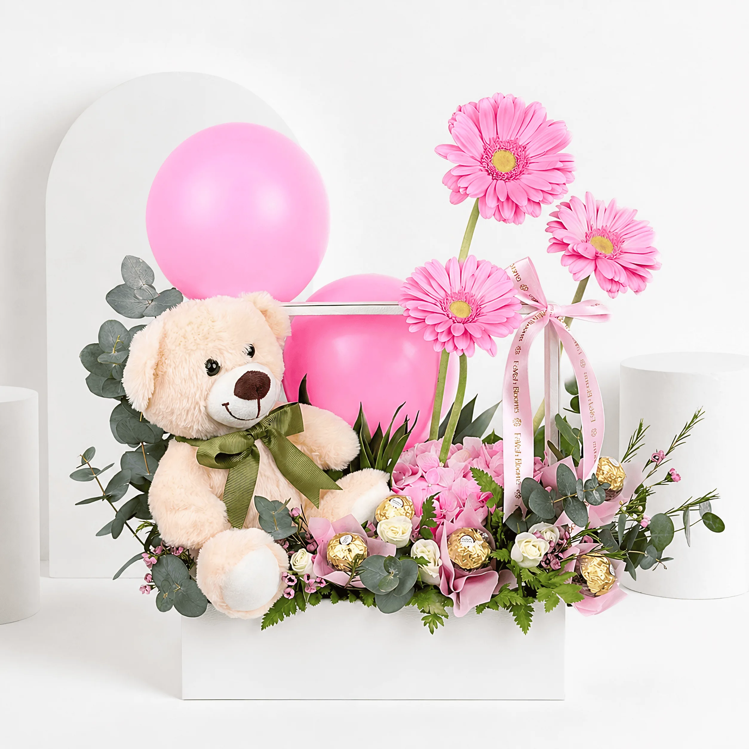 Luxury pink gift arrangement with teddy bear, pink balloons, gerbera flowers, Ferrero Rocher chocolates, eucalyptus, and fresh greenery in a white box on a white studio background.