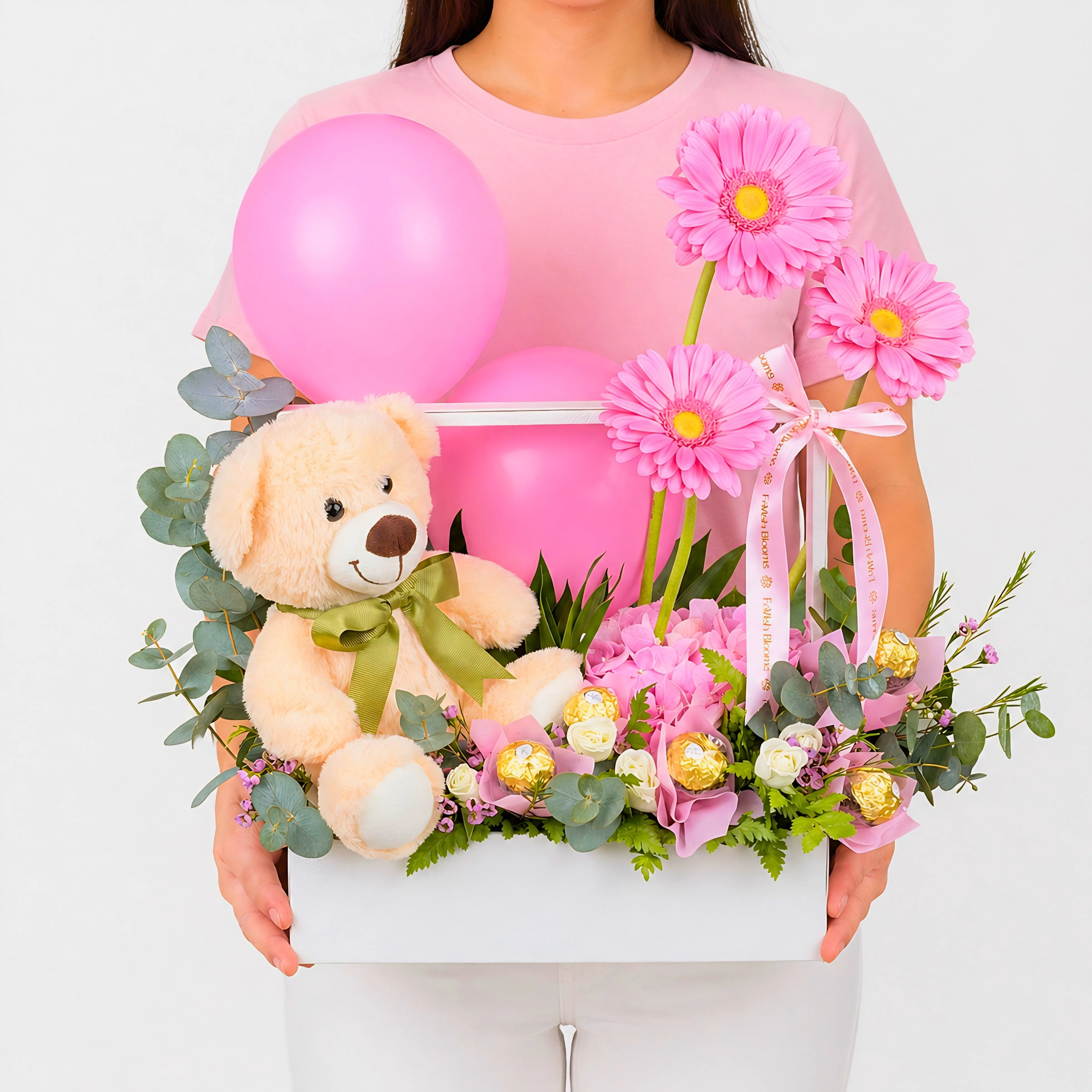 Woman holding a luxury pink gift box with teddy bear, pink balloons, gerbera flowers, Ferrero Rocher chocolates, and fresh floral greenery.
