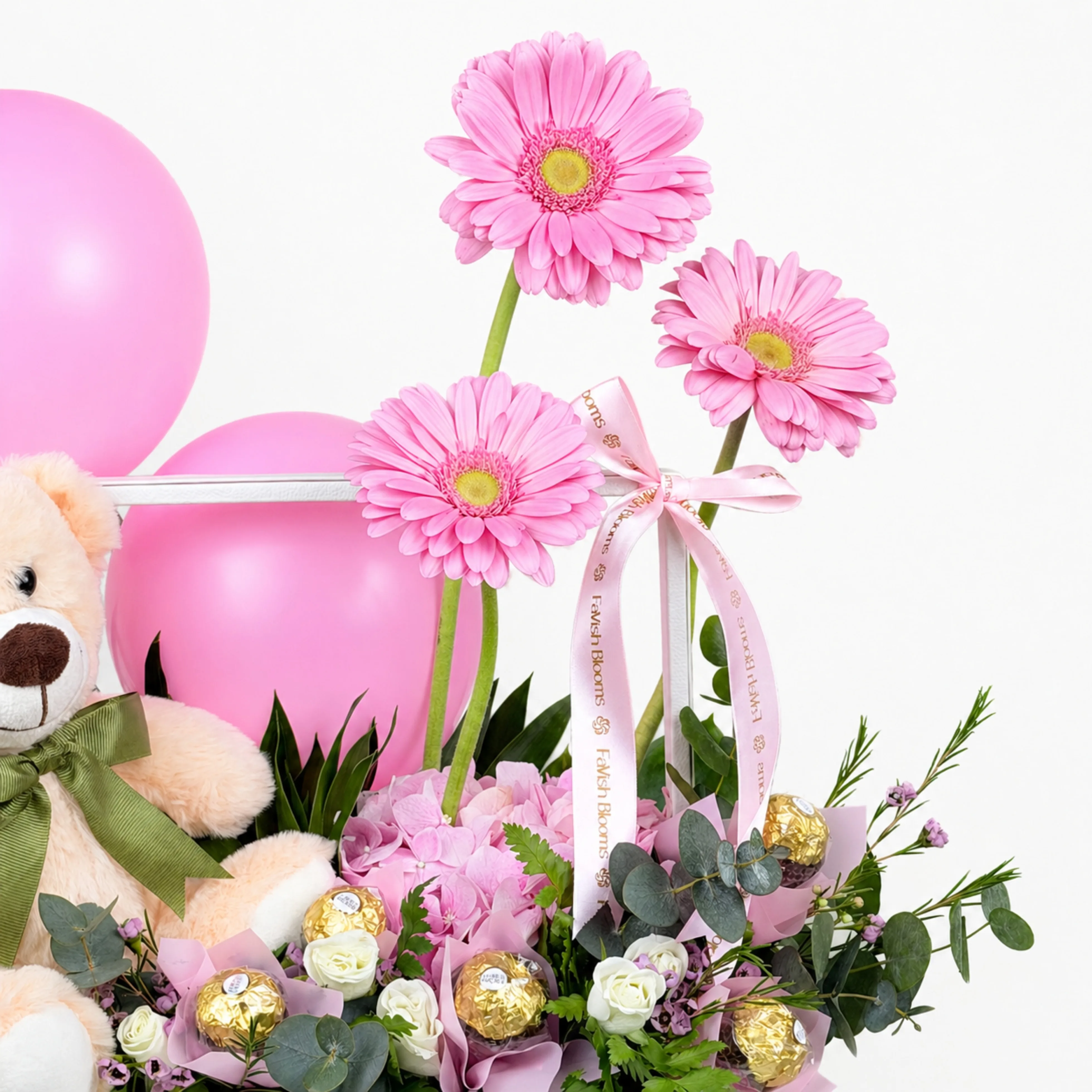 Pink floral gift arrangement with gerbera flowers, balloons, teddy bear, Ferrero Rocher chocolates, ribbon, eucalyptus, and soft greenery.