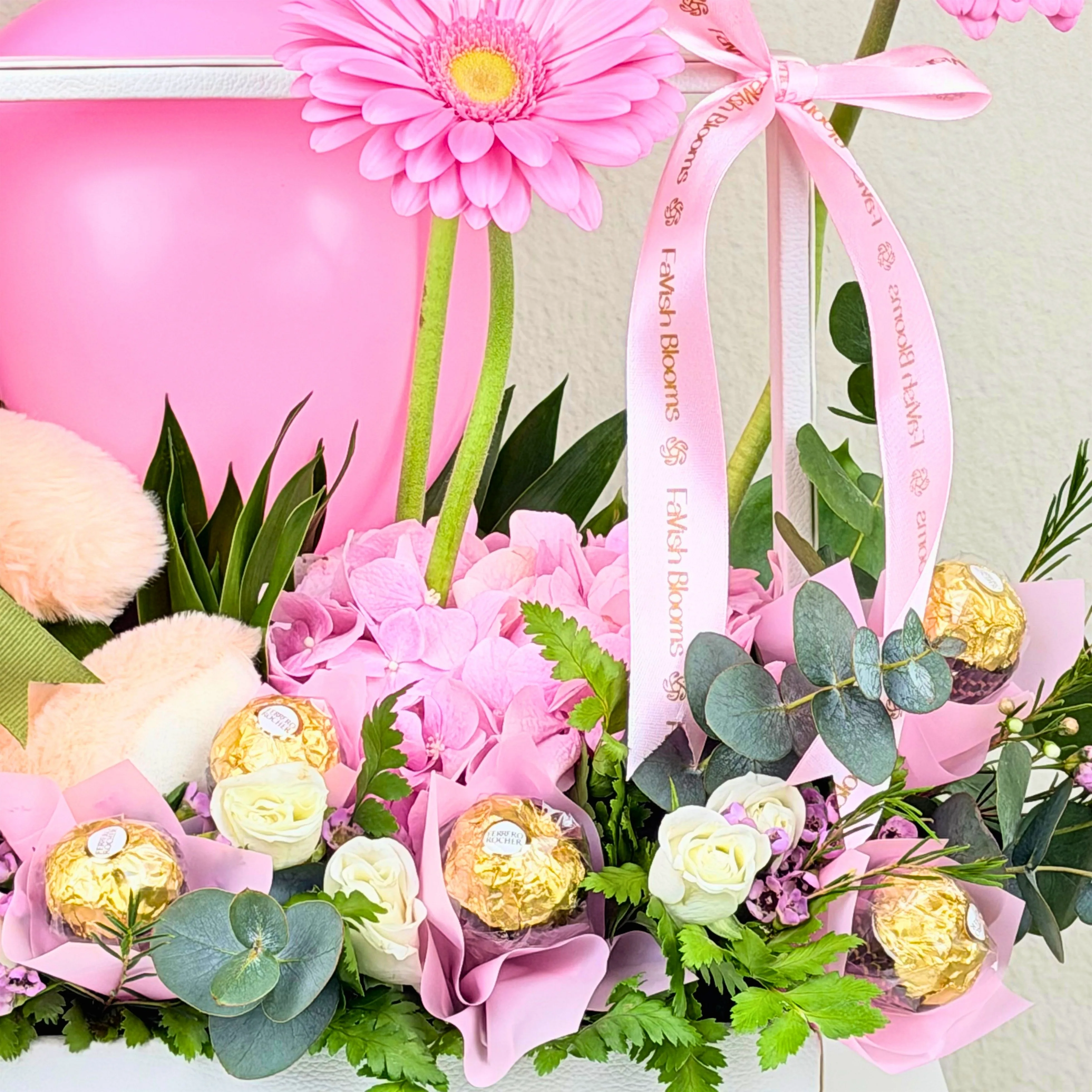 Close-up of pink hydrangea and gerbera gift arrangement with Ferrero Rocher chocolates, white roses, eucalyptus, ribbon, and fresh greenery.