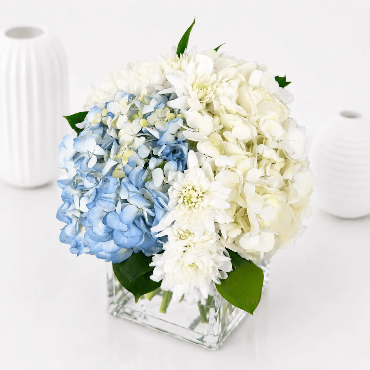 Blue and white hydrangea glass vase arrangement with fresh white chrysanthemums and green leaves.