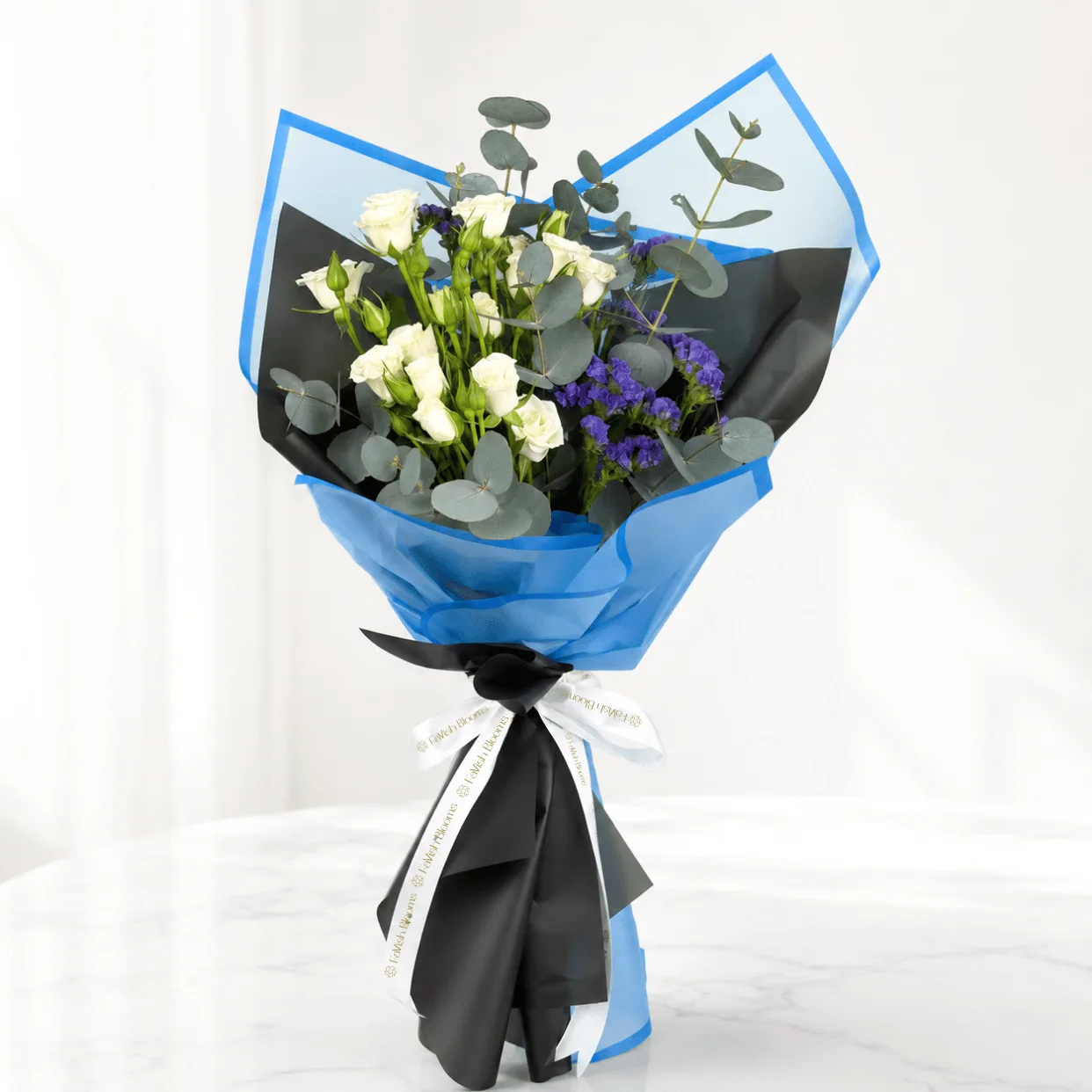 Blue wrapped white rose bouquet with purple statice, eucalyptus and black ribbon on a marble table.