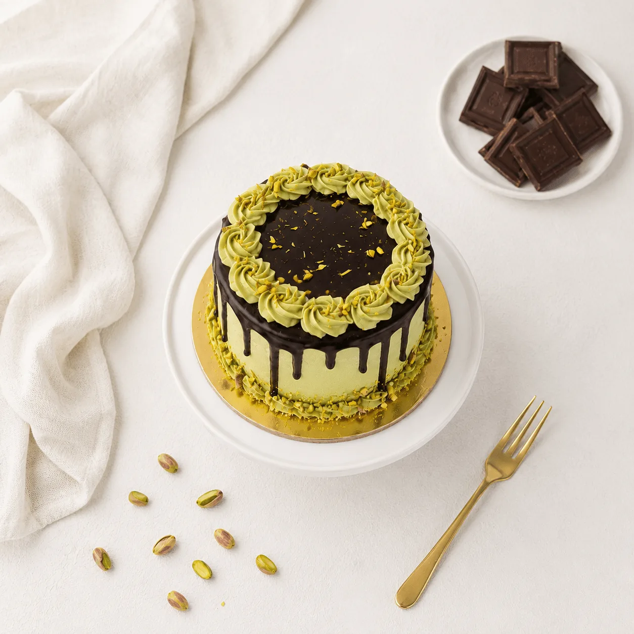 "Overhead view of a pistachio cake with green frosting rosettes and chocolate ganache drip, displayed on a white cake stand alongside a gold fork, scattered pistachios, chocolate squares, and a linen cloth."