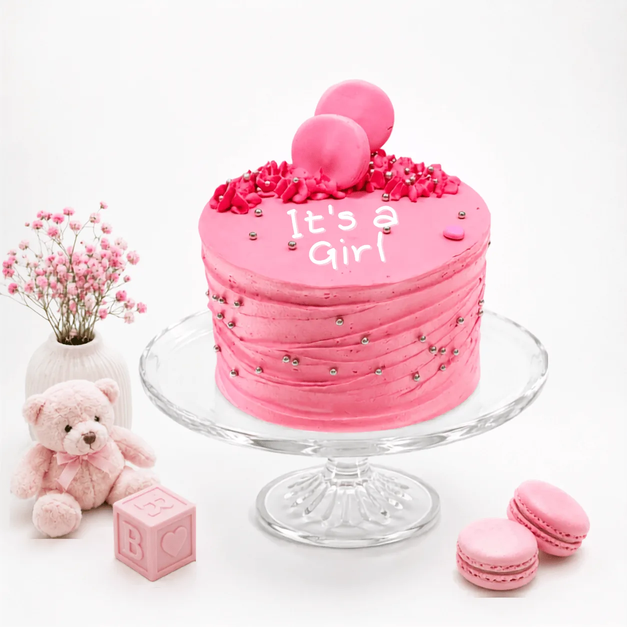 "Pink 'It's a Girl' cake with silver pearls and macaron toppers, surrounded by baby shower decor."