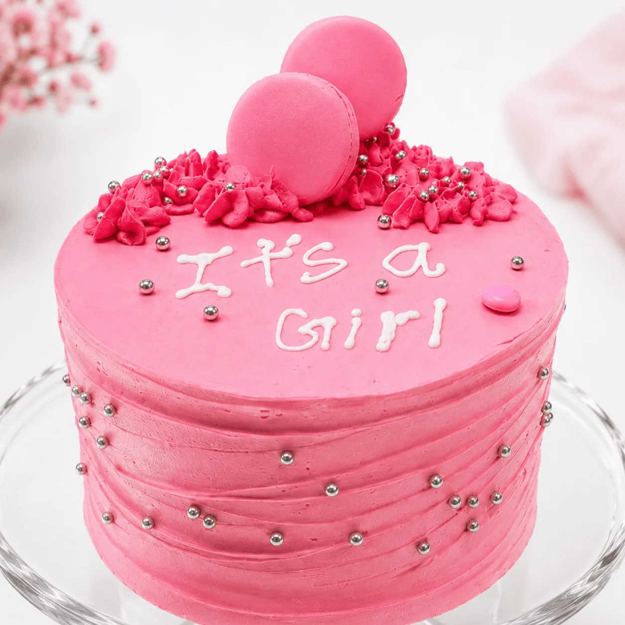 Close-up of pink "It's a Girl" cake with macaron toppers and silver edible pearls.