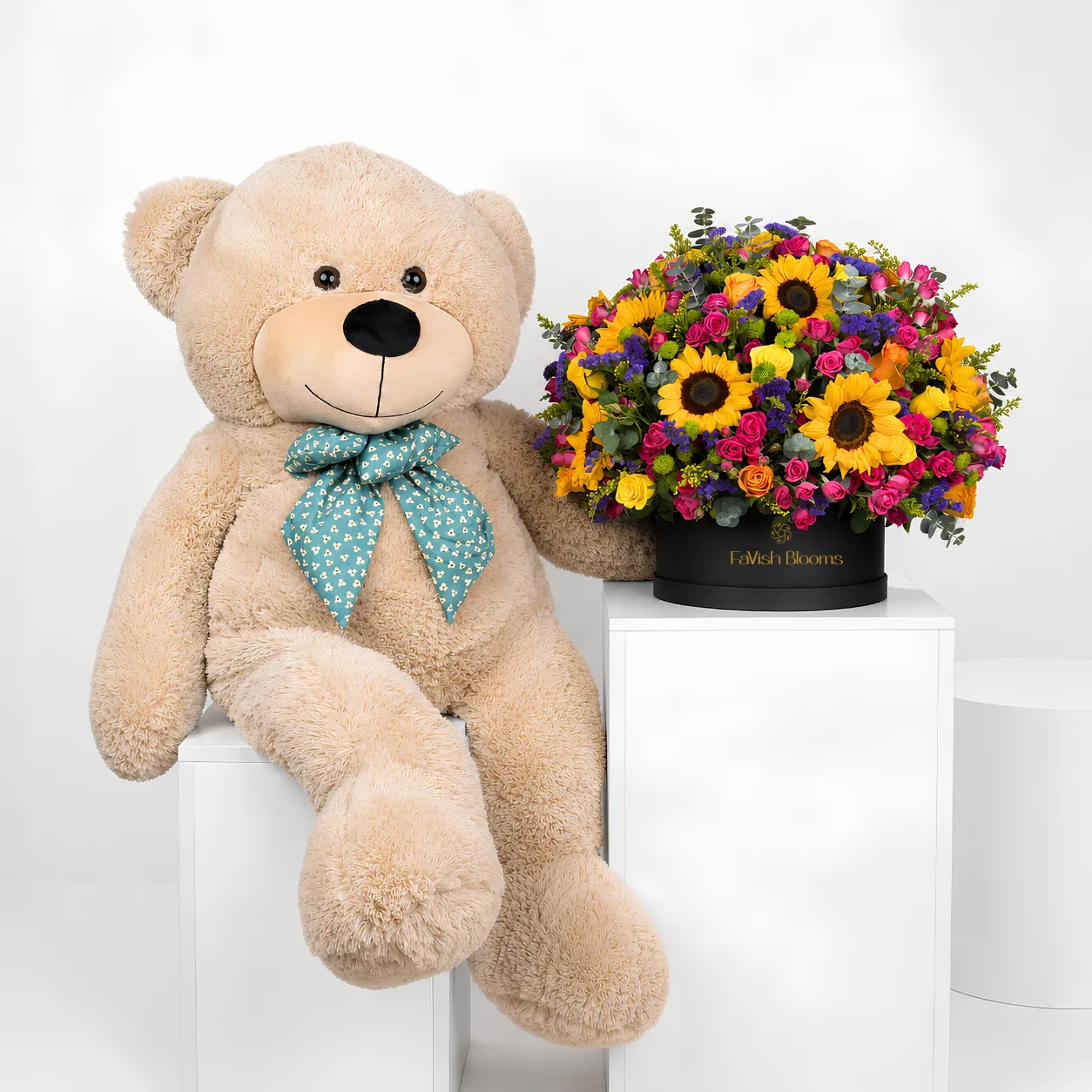 "A large beige teddy bear with a teal bow sits beside a colorful mixed flower arrangement of sunflowers, roses, and greenery in a black 'FaVish Blooms' hat box, both displayed on white pedestals."