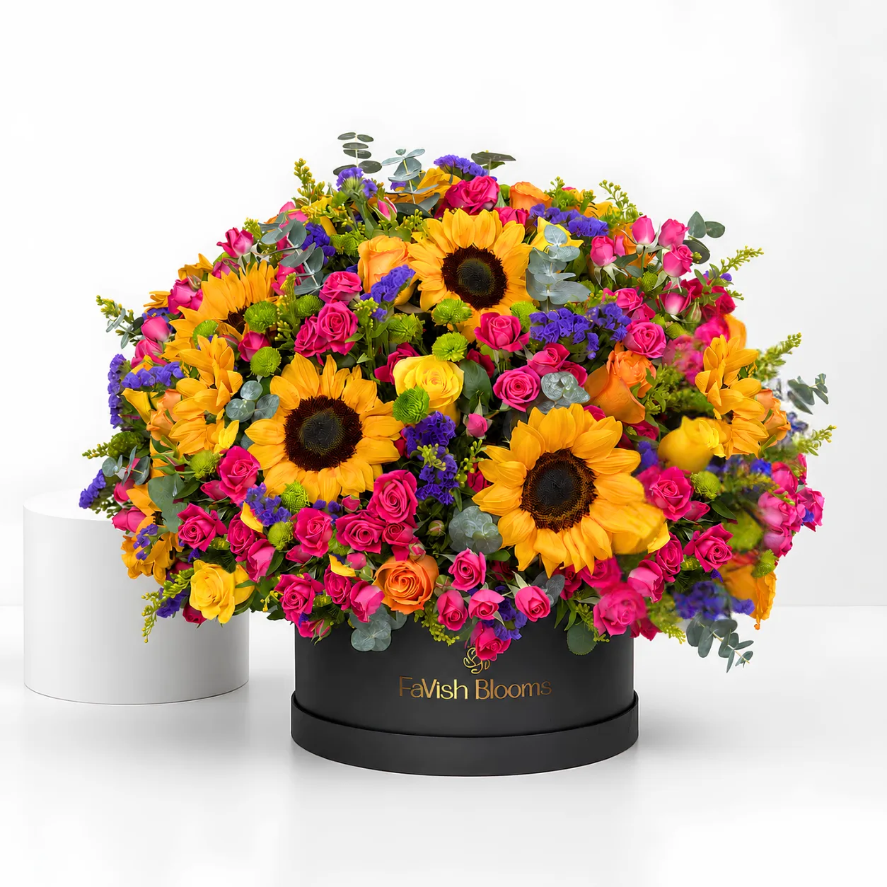 "A vibrant flower arrangement of sunflowers, pink and orange roses, purple statice, and eucalyptus in a black 'FaVish Blooms' hat box, displayed on a white table."