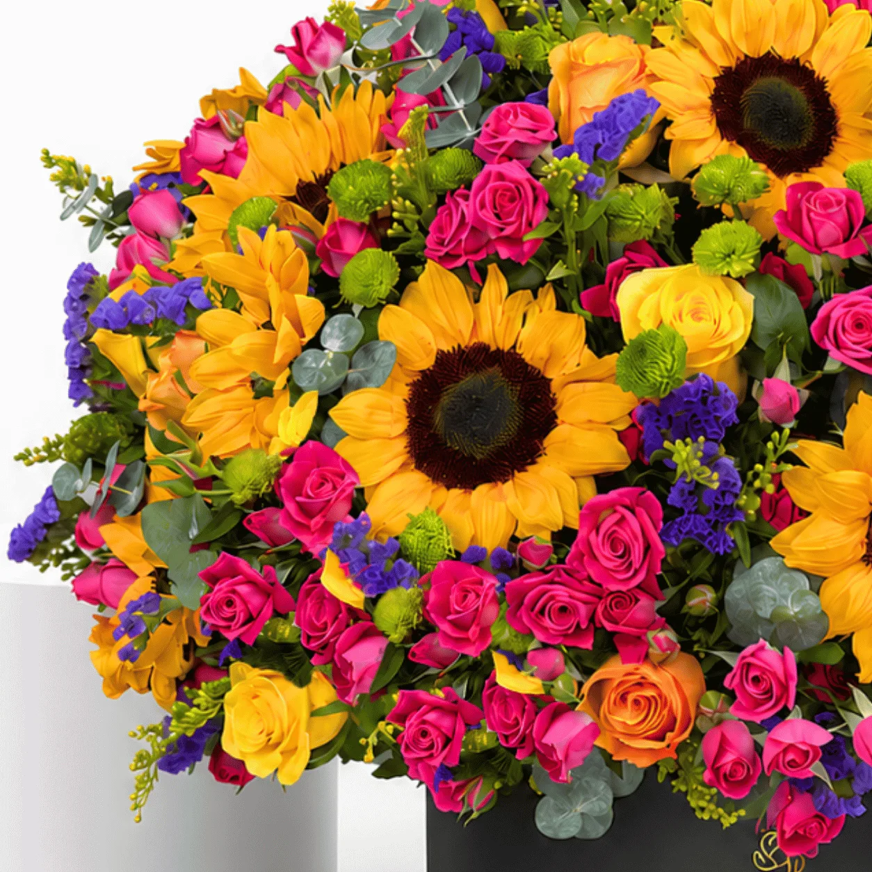 "Close-up of a colorful flower arrangement with sunflowers, pink and orange roses, purple statice, and green button chrysanthemums, arranged in a black hat box."