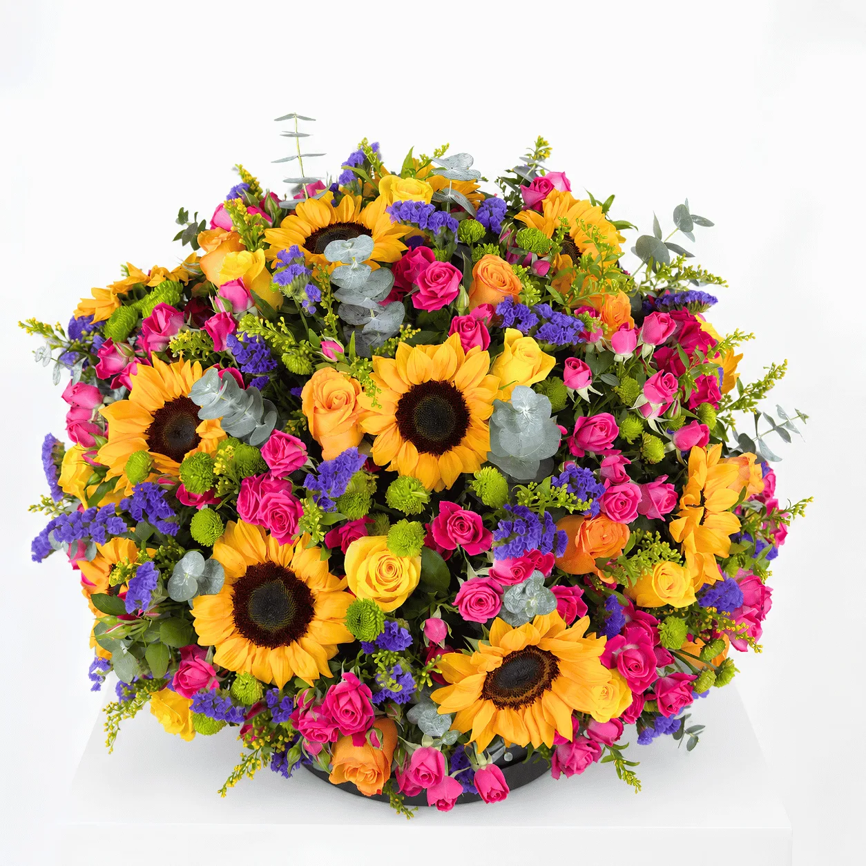 "A lush, colorful flower arrangement featuring sunflowers, pink and orange roses, purple statice, green button chrysanthemums, and eucalyptus, arranged in a round black hat box on a white surface."