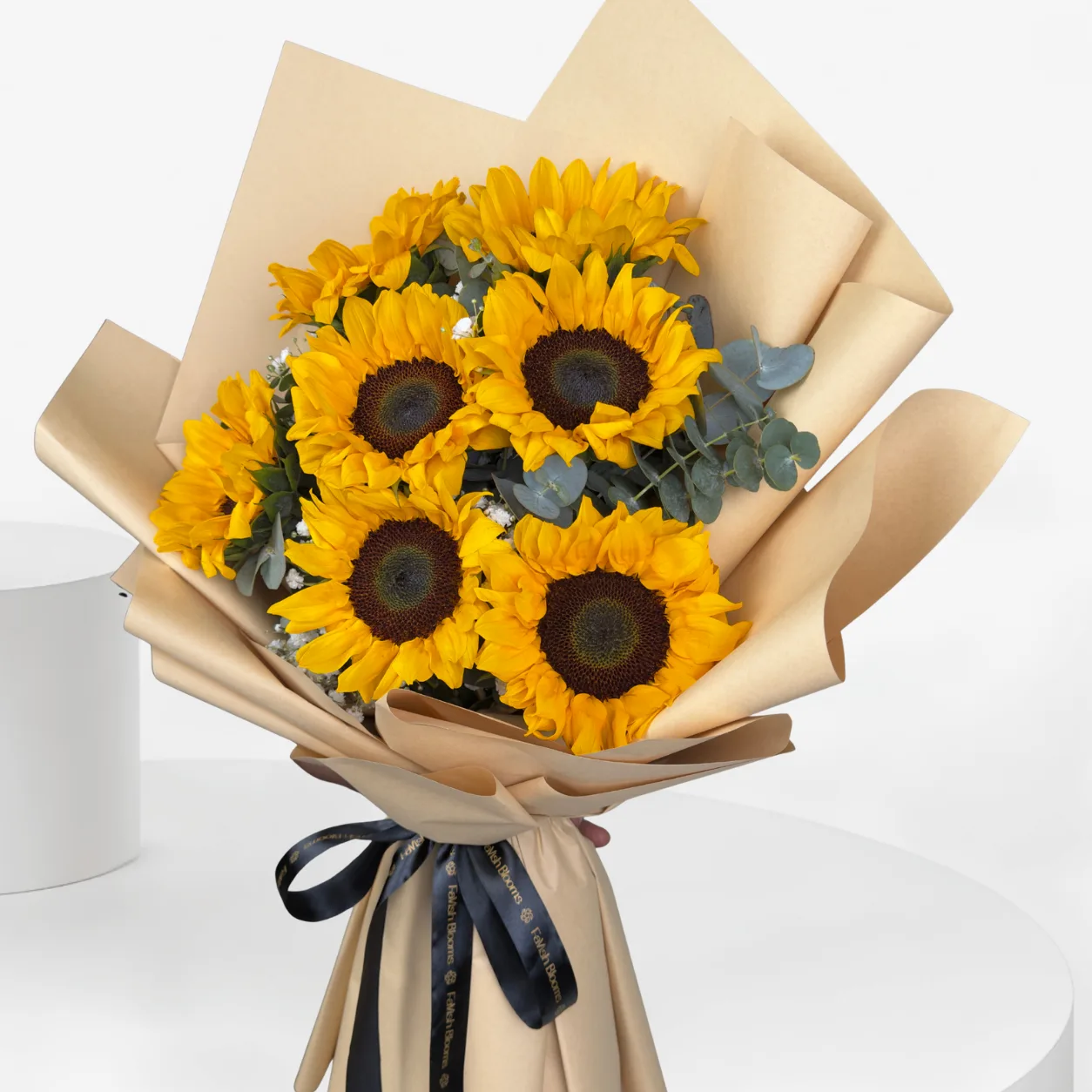 "A hand holds a sunflower bouquet wrapped in beige kraft paper with eucalyptus greenery, tied with a black ribbon, set against a white pedestal backdrop."